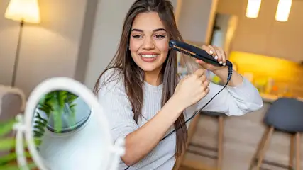 A smiling woman straightens her long hair with a flat iron at home, looking into a mirror in warm, soft lighting.