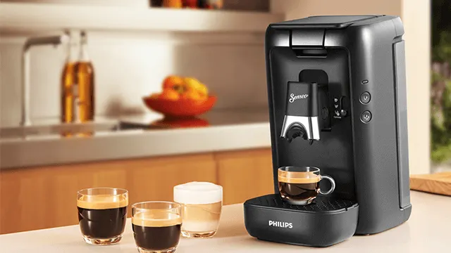 Een zwarte Senseo koffiemachine met drie glazen: twee met koffie, één met melk. Op de achtergrond een keuken.