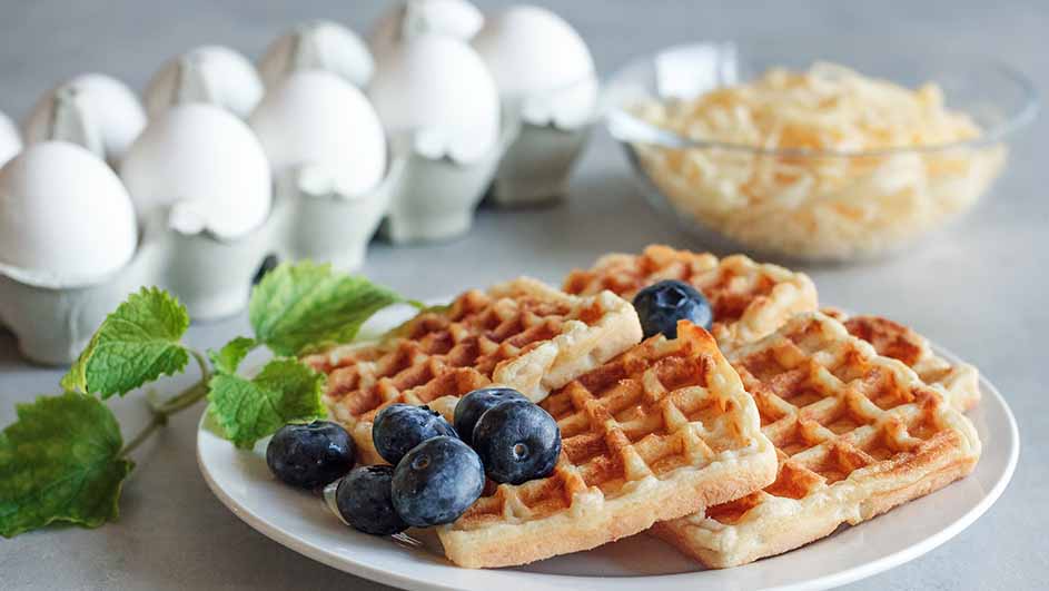 Wafels met blauwe bessen en groene blaadjes op een wit bord, eieren in een houder en een kom met geraspte kaas op de achtergrond.