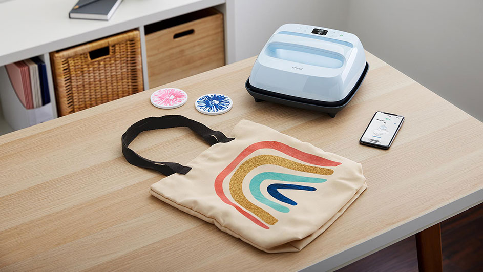 A tan tote bag with a rainbow design sits on a wooden table. Beside it is a white heat press, a phone, and two coasters.