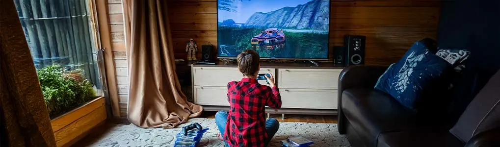 Een kind zit op de grond en speelt een racespel op een gameconsole, zichtbaar op een groot tv-scherm. Voor hem liggen stapels gamehoesjes en een extra controller.
