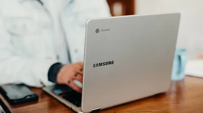 Een zilverkleurige Samsung-laptop staat op een houten tafel, met een telefoon en een blauwe mok op de achtergrond.