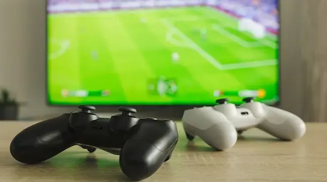 Two game controllers, black and white, sit on a wooden surface in front of a TV screen showing a soccer game.