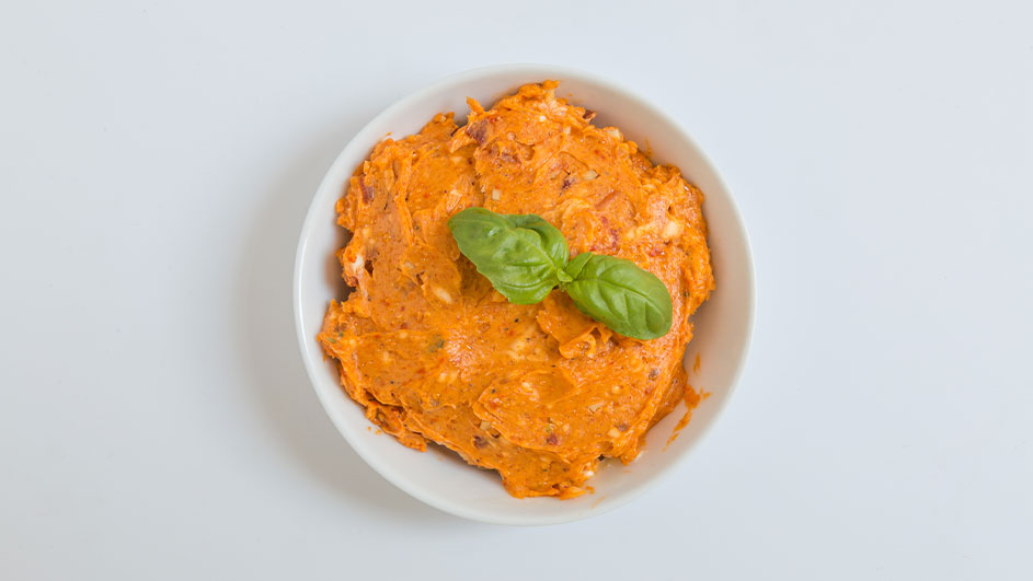 Oranje smeersel in een wit schaaltje, gegarneerd met basilicumblaadjes, op een witte achtergrond.