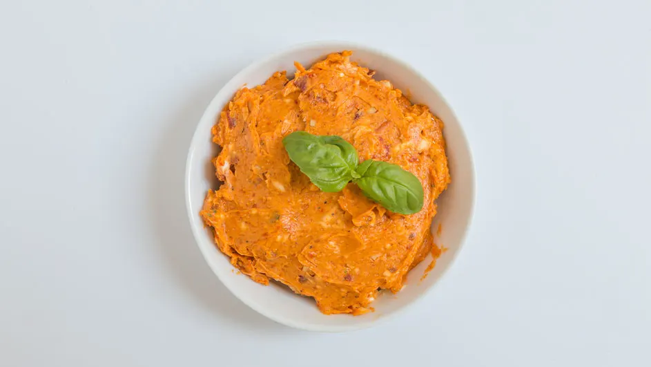 Oranje smeersel in een wit schaaltje, gegarneerd met basilicumblaadjes, op een witte achtergrond.