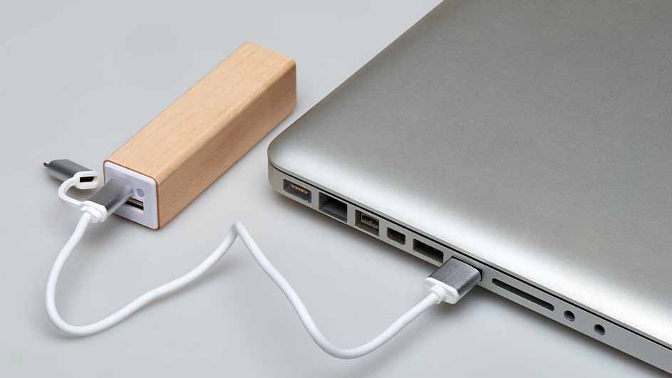 Een zilverkleurige laptop is met een witte kabel verbonden met een rechthoekige, bruine powerbank op een grijze ondergrond.