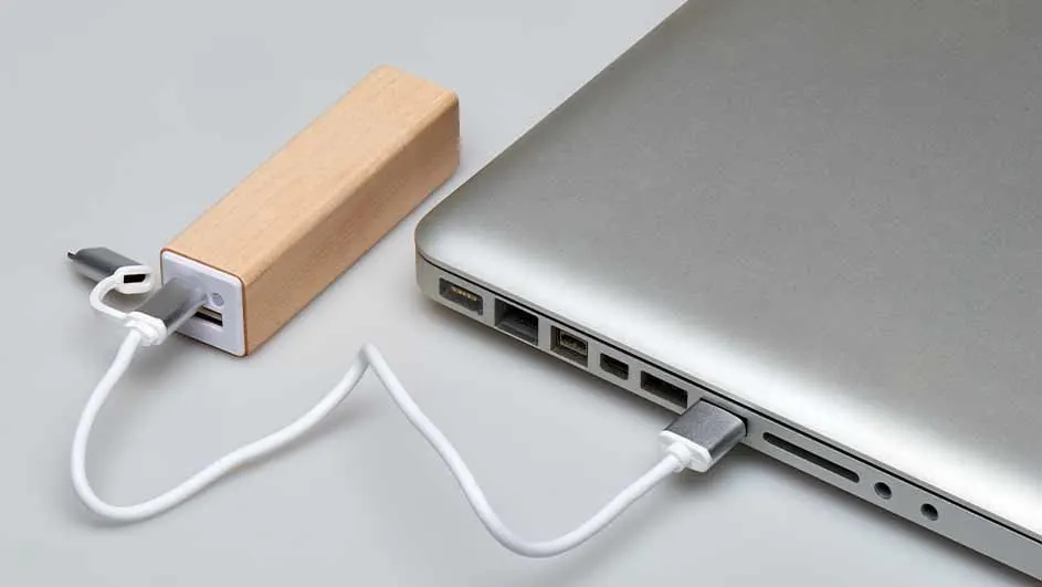 Een zilverkleurige laptop is met een witte kabel verbonden met een rechthoekige, bruine powerbank op een grijze ondergrond.