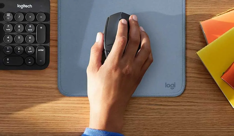 Hand holding a black mouse on a blue Logitech mat, next to a compact keyboard and colourful folders on a wooden desk.