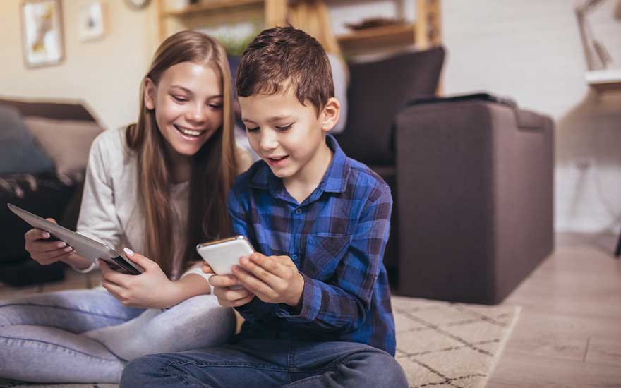 Two children sit relaxed on the floor, smiling as they play together on a smartphone and a tablet in the living room at home.
