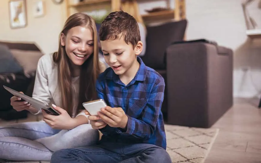 Two children sit relaxed on the floor, smiling as they play together on a smartphone and a tablet in the living room at home.