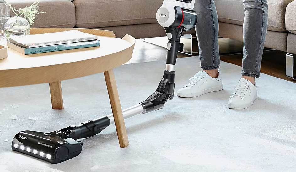 A person hoovers a bright living room with a sleek, flexible Bosch cordless vacuum under a wooden coffee table on a soft rug.