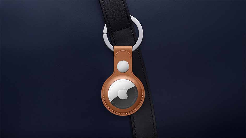 Een blauwe tas met een zwarte riem waaraan een Apple AirTag hangt met een bruine lederen houder en een zilveren ring.