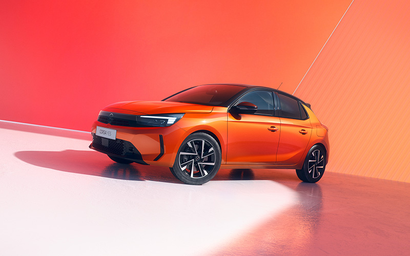 Oranje Opel Corsa-e geparkeerd voor een rode achtergrond, zijaanzicht met moderne velgen en strakke lijnen.