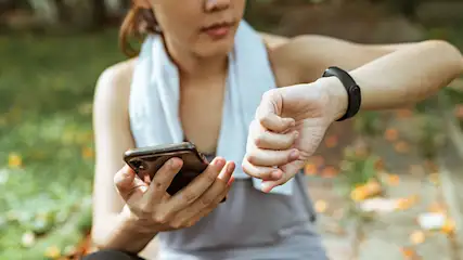 Een vrouw in sportkleding kijkt naar een sporthorloge en houdt een telefoon vast, met een witte handdoek om haar nek en een groenachtige achtergrond.