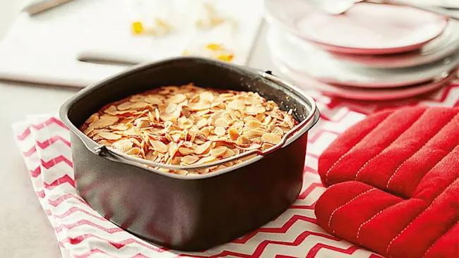 Een vierkante bakvorm gevuld met amandelschilfers staat op een rood-wit gestreept doek, naast een rode ovenwant en borden.