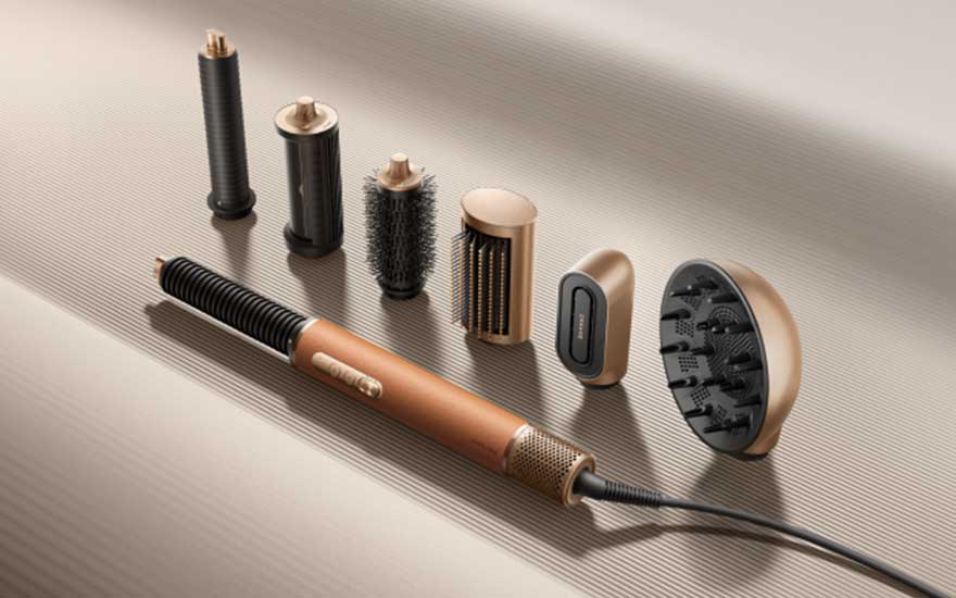 Een koperkleurige haarstyler ligt met meerdere opzetstukken, borstels en diffuser op een geribbeld beige oppervlak.