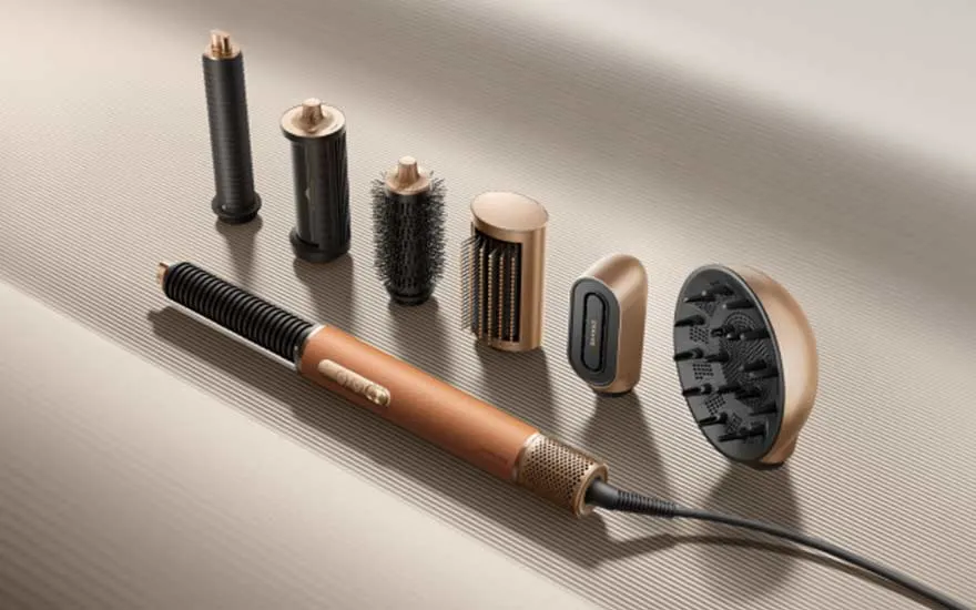 Een koperkleurige haarstyler ligt met meerdere opzetstukken, borstels en diffuser op een geribbeld beige oppervlak.