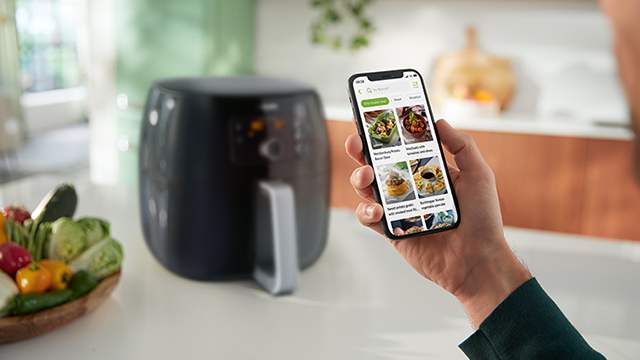 Een zwarte airfryer staat op een aanrecht naast een houten schaal met groenten. Een hand houdt een telefoon met een recepten-app.