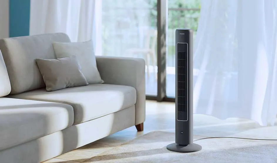 A Philips tower fan stands by a light sofa in a bright living room with curtains and a large window.