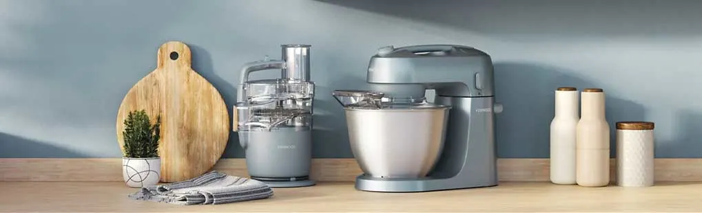 Op een houten aanrecht staan een grijze keukenmachine en blender; ernaast snijplank, doek, plantje en twee flessen tegen een blauwe muur.