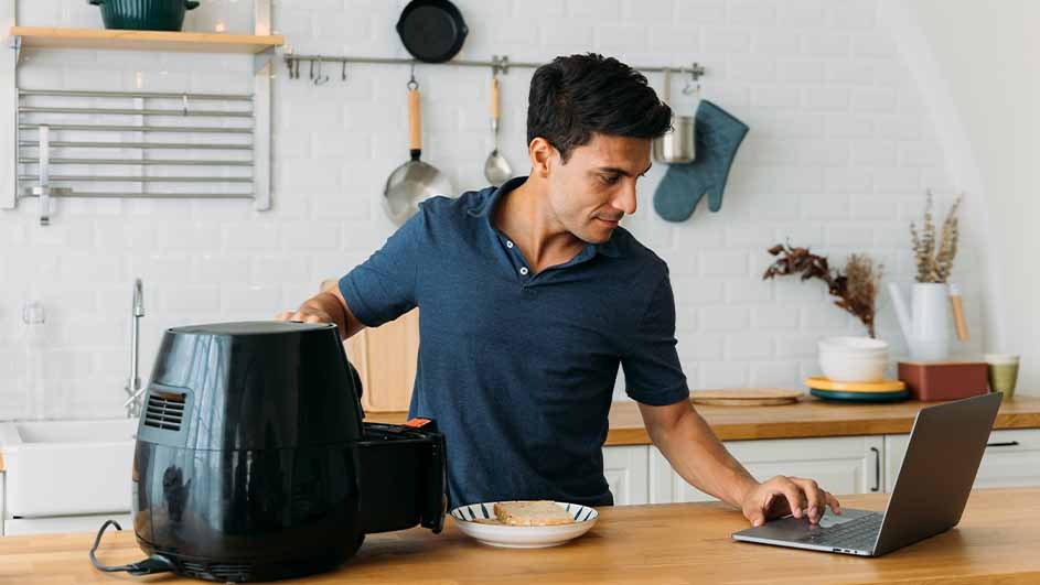 A person in a blue shirt uses a laptop next to a black air fryer on a wooden counter in a kitchen.