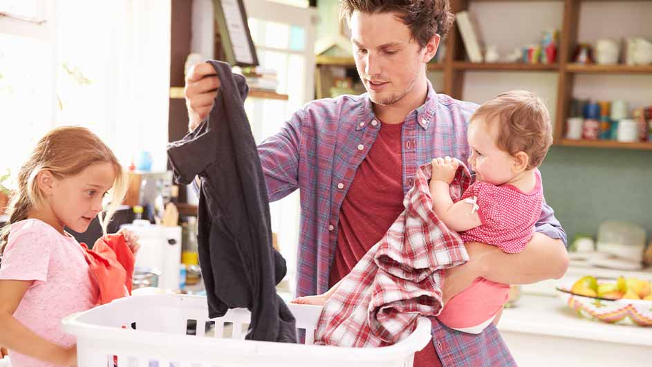 Een vader met baby en dochter vouwt wasgoed boven een witte mand in een huiselijke setting.