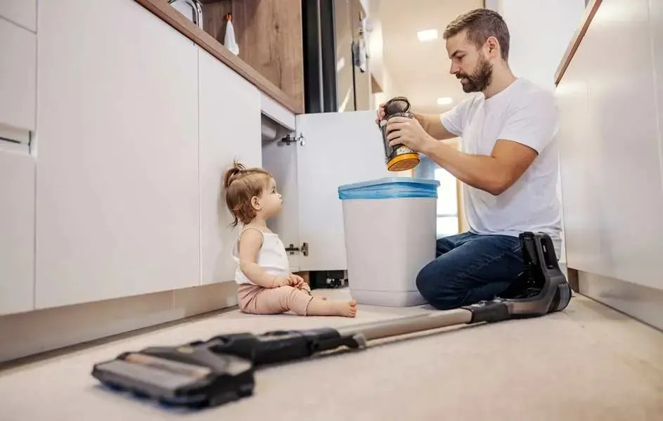 A man empties a stick vacuum cleaner’s dust container into a bin, while a small child sits on the floor in a modern kitchen.