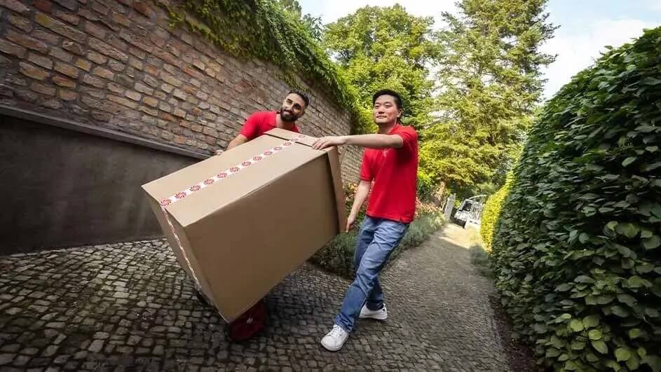 Twee mensen in rode shirts vervoeren samen een groot kartonnen Bosch-doos over een stenen pad omgeven door groen.