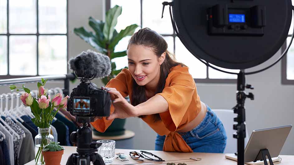 Een vrouw in een oranje blouse filmt met een camera op een statief voor een raam en een lamp. Er staan tulpen en accessoires op tafel.