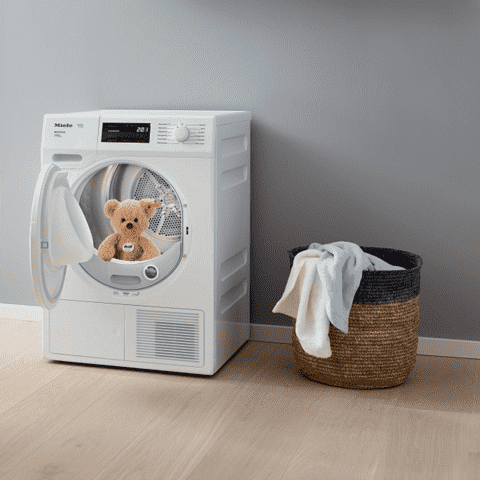 Witte droger met open deur en teddybeer erin. Naast de droger staat een gevlochten wasmand met witte was.