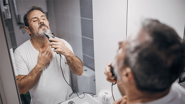 Man scheert baard in de badkamer voor de spiegel met een zwarte tondeuse en een wit T-shirt.