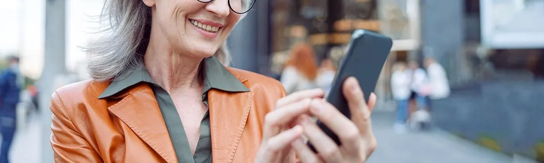 A smiling woman wearing glasses and an orange jacket is outdoors, holding and using a smartphone. No visible brand appears on the phone or clothing.