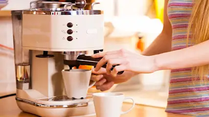 Een vrouw bedient een crèmekleurig koffiezetapparaat met twee witte mokken eronder en ervoor.