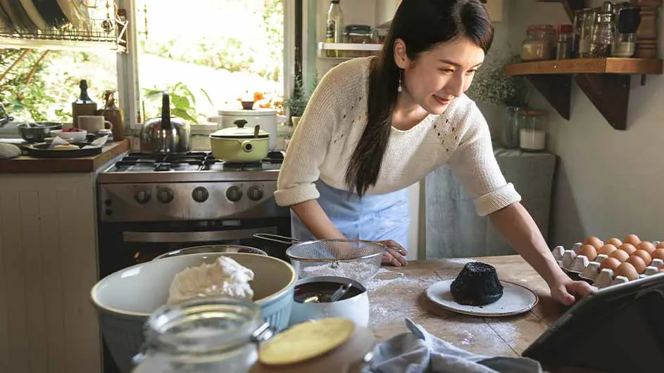Een vrouw bakt in een keuken. Er staat een fornuis, een aanrecht met ingrediënten en een bakvorm.