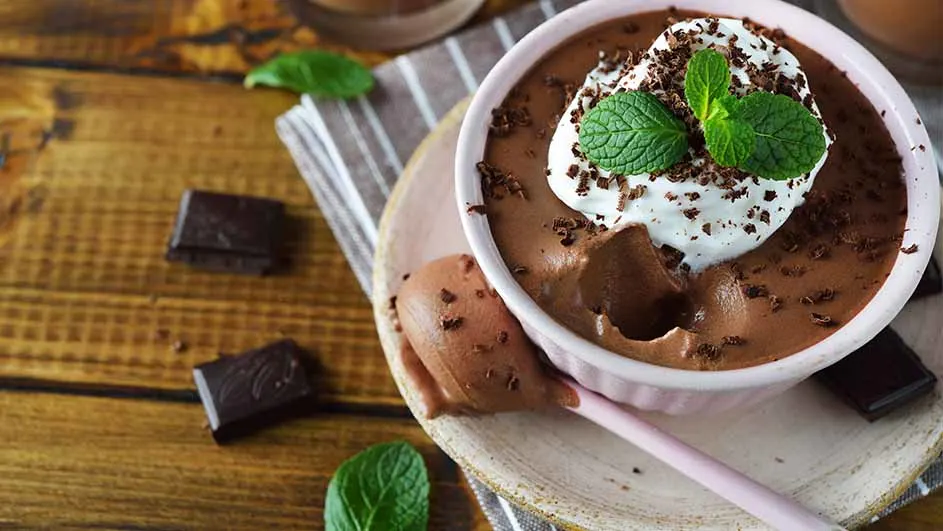Chocoladepudding in een roze kom met slagroom en munt, op een houten ondergrond met chocolade en een gestreepte doek.