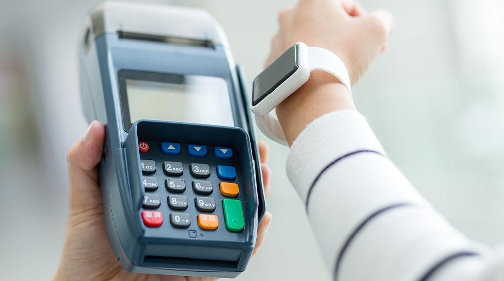 Een hand houdt een blauw betaalautomaat vast met kleurrijke knoppen, naast een pols met een witte smartwatch.