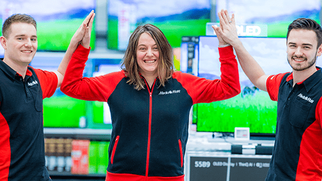 Drie lachende mensen in zwarte en rode MediaMarkt-kleding geven elkaar een high-five voor een achtergrond van televisies in een winkel.