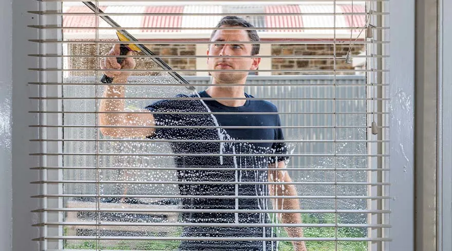 Een man reinigt een raam met witte jaloezieën en een wisser; zeepresten zichtbaar. Buiten een hek en een bakstenen gebouw.