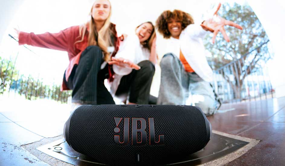 Drie vrolijke vrienden poseren buiten achter een zwarte JBL-bluetoothspeaker op de voorgrond, armen uitgestrekt naar de camera.