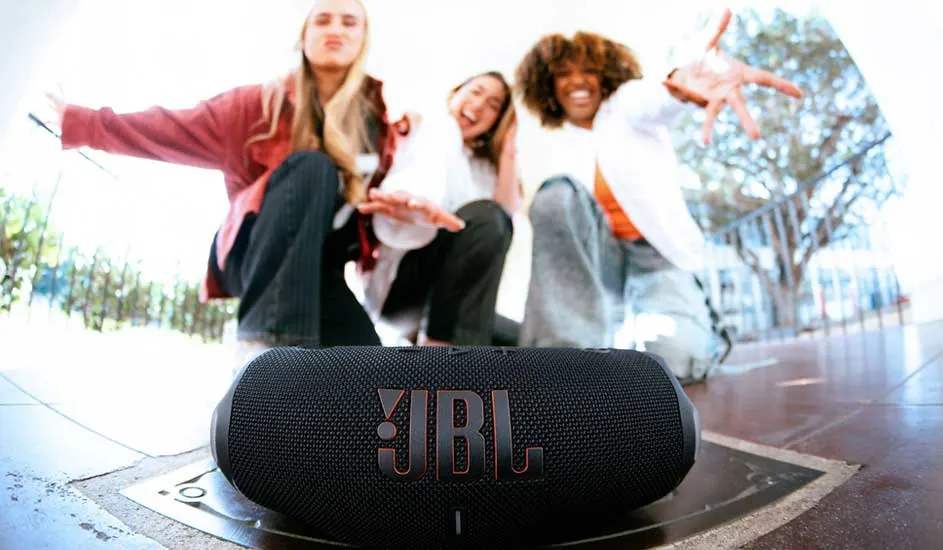 Drie vrolijke vrienden poseren buiten achter een zwarte JBL-bluetoothspeaker op de voorgrond, armen uitgestrekt naar de camera.