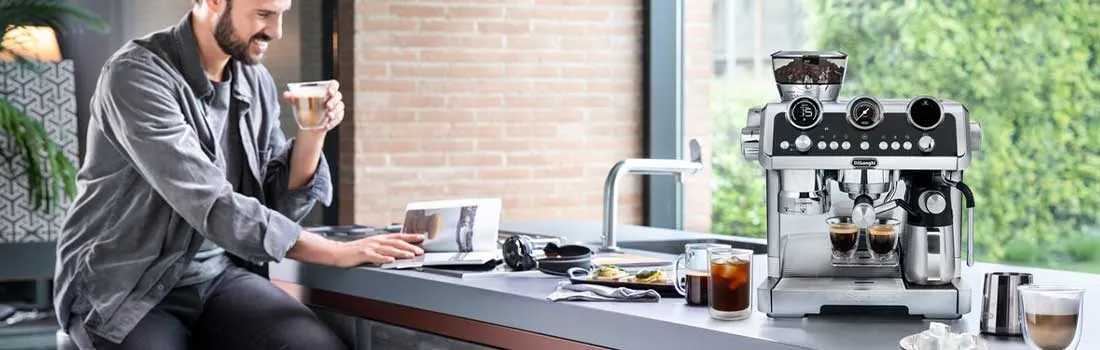 A smiling man works at the kitchen counter with his laptop and coffee, while a large silver espresso machine stands beside him.