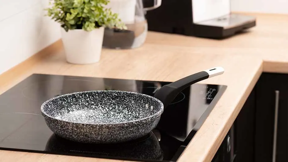 Een grijze koekenpan met zwart handvat op een zwarte kookplaat. Op de achtergrond een plant, een waterkan en een koffiezetapparaat.