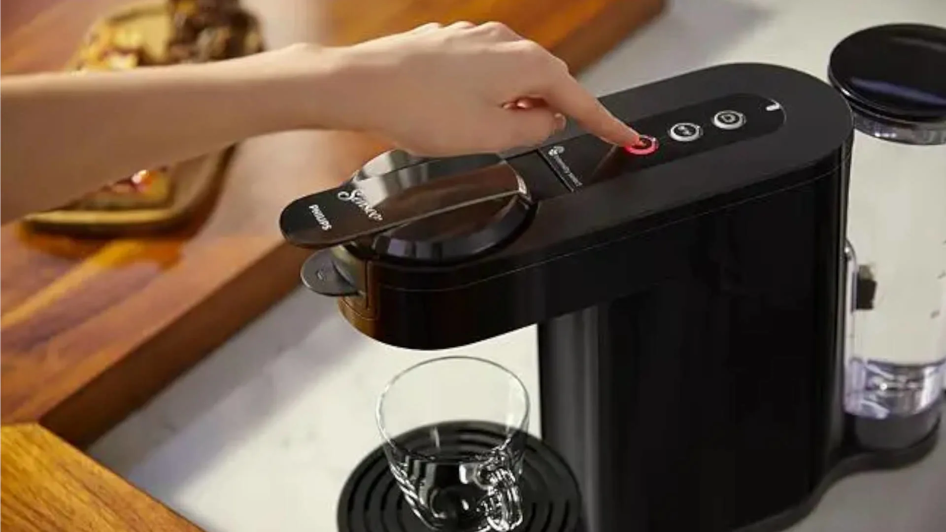 Een zwarte Senseo koffiemachine met een hand die een rode knop indrukt en een kopje eronder.