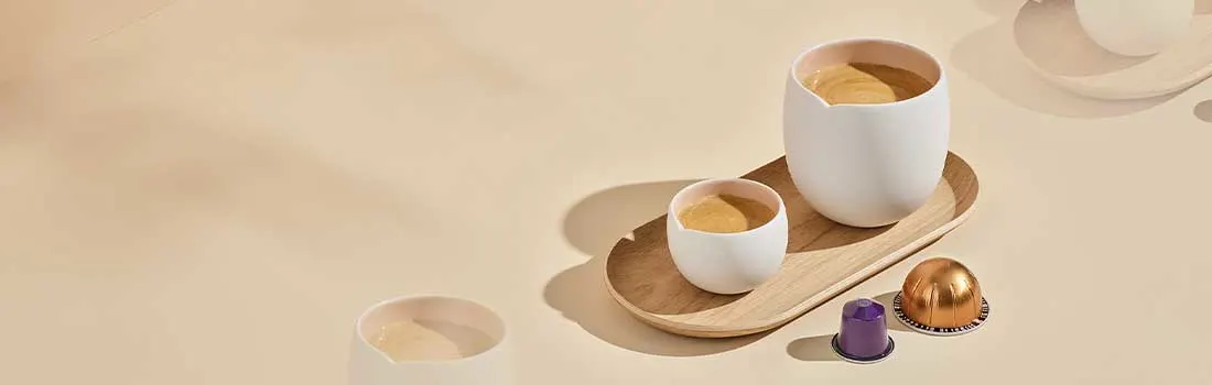 Twee koffiekopjes met romige espresso staan op een houten dienblad, naast een paar koffiecapsules op een lichte achtergrond.