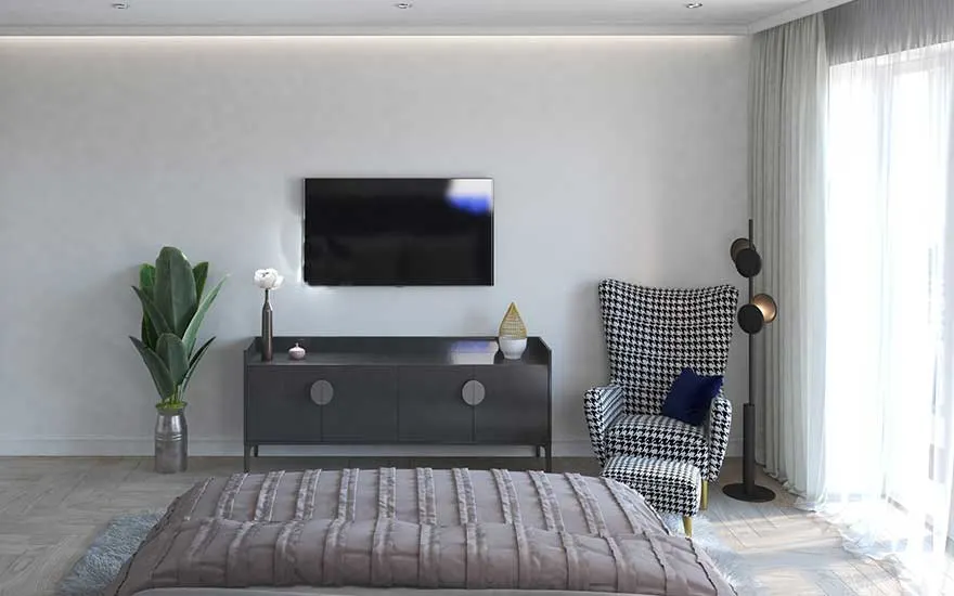 In a bright bedroom, a TV is mounted on the wall across from the bed, with an armchair, plants, and cabinet nearby.