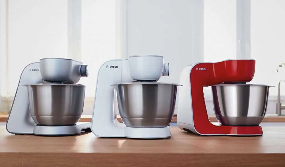 Drie Bosch keukenmachines met metalen kommen staan op een houten aanrecht: twee wit/grijs en één rood, voor een helder raam in de keuken.