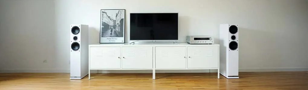 Witte kast met tv, audiospeler, zwart-wit foto en twee witte luidsprekers op een houten vloer tegen een witte muur.