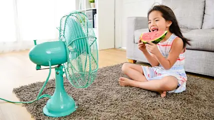 Meisje eet watermeloen naast een turquoise ventilator op een bruin tapijt in een kamer met een grijze bank.