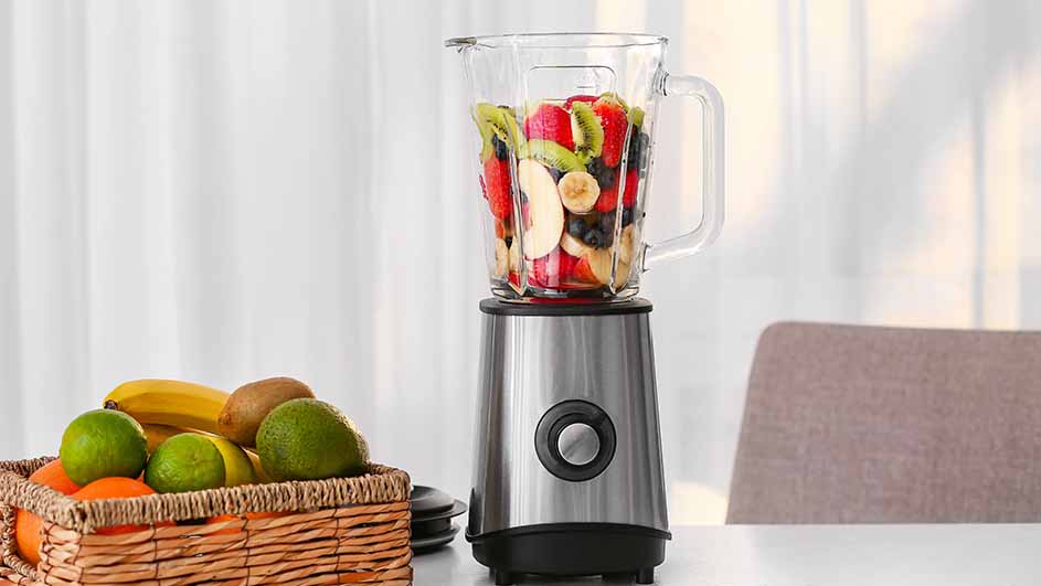 Een blender met fruit staat op een tafel. Naast de blender staat een mand met bananen, kiwi's en limoenen.