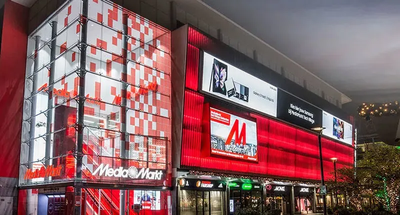 Winkelpand Media Markt met rode en witte decoratie en reclameborden. Winkels op de begane grond met verlichting.